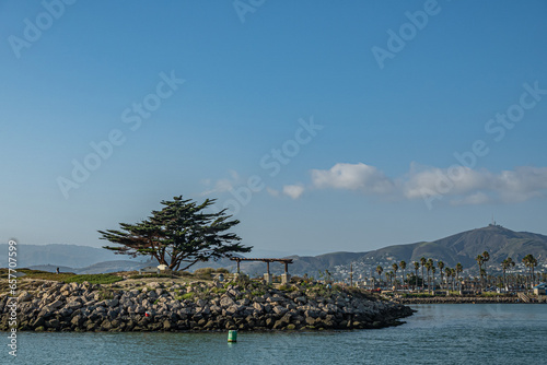 Ventura, CA, USA - September 14, 2023: Soter's Point Marina Park tree and benches above rocks forming pier under blue sky. Mountain range and downtown in back. photo