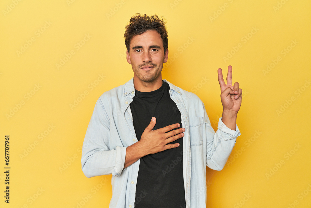 Young Latino man posing on yellow background taking an oath, putting hand on chest. Young Latino man posing on yellow background taking an oath, putting hand on chest.