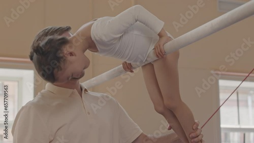 Medium shot of Caucasian male trainer helping 5 year old Asian girl to do pullover on gymnastics bar during indoor training