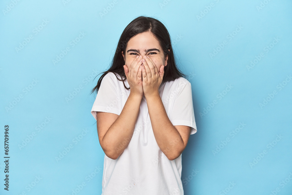 Young Caucasian woman on blue backdrop laughing about something, covering mouth with hands. Young Caucasian woman on blue backdrop laughing about something, covering mouth with hands.