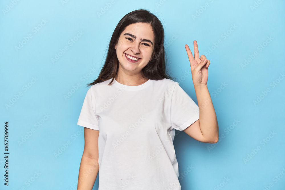 Young Caucasian woman on blue backdrop joyful and carefree showing a peace symbol with fingers. Young Caucasian woman on blue backdrop joyful and carefree showing a peace symbol with fingers.