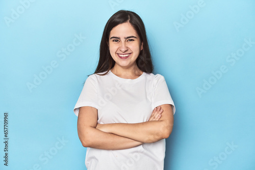 Young Caucasian woman on blue backdrop who feels confident, crossing arms with determination.