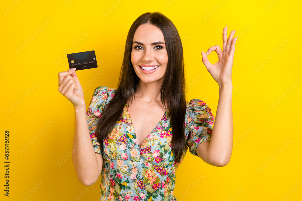 Portrait of satisfied clever woman with long hairstyle wear stylish dress hold debit card show okey isolated on yellow color background Portrait of satisfied clever woman with long hairstyle wear stylish dress hold debit card show okey isolated on yellow color background