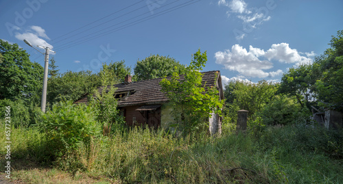 Stary opuszczony dom z zapadłym dachem stojący wśród bujnych drzew pod błękitnym niebem z białymi chmurkami. Lato w lesie.