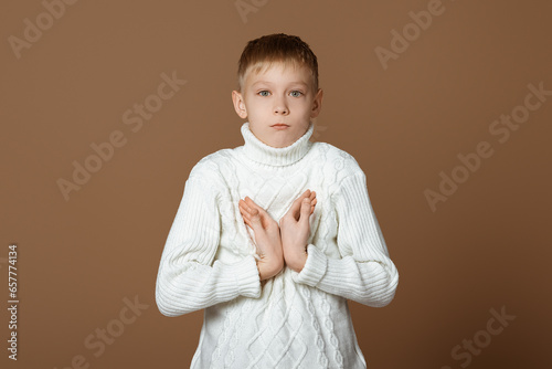 Shocked and worried teen boy pointing fingers at himself, frowning and sulking upset, standing in white knitted sweater against beige background
