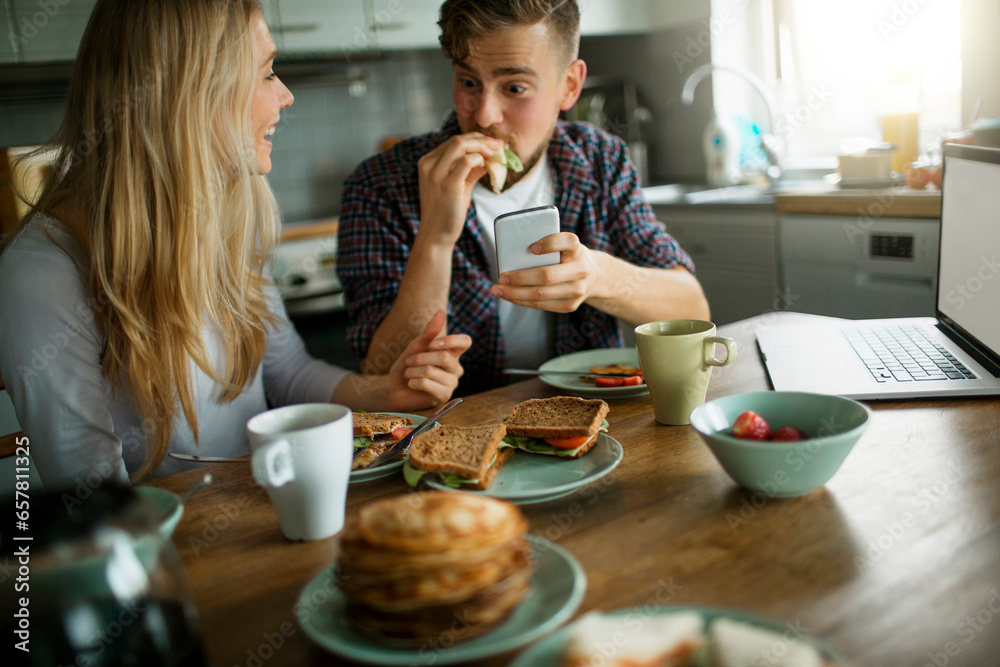 Young couple using a smartphone while having breakfast together in the morning in the kitchen at home Young couple using a smartphone while having breakfast together in the morning in the kitchen at home