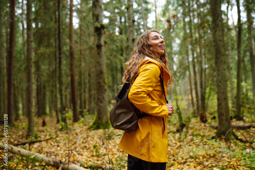 Happy woman in a yellow coat enjoys the green beautiful natural forest around her. A tourist with a backpack is exploring new places, walking through the thicket of the forest.