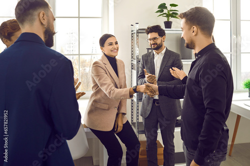 Team of business people congratulating young woman on her success. Happy young woman smiles and shakes hands with man while colleagues are applauding. Recognition at work concept