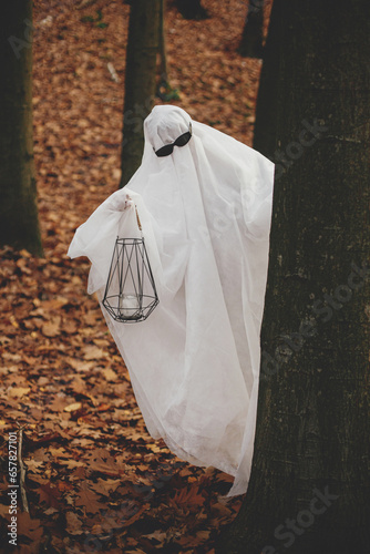 Funny ghost in black glasses holding lantern and peeking out of a tree in autumn forest. Person dressed in white sheet as stylish ghost trick or treating in evening fall woods. Happy Halloween