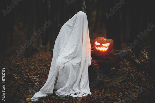 Happy Halloween! Spooky ghost sitting with glowing jack o lantern in moody dark autumn forest. Person dressed in white sheet as ghost with pumpkin in evening fall woods. Boo!