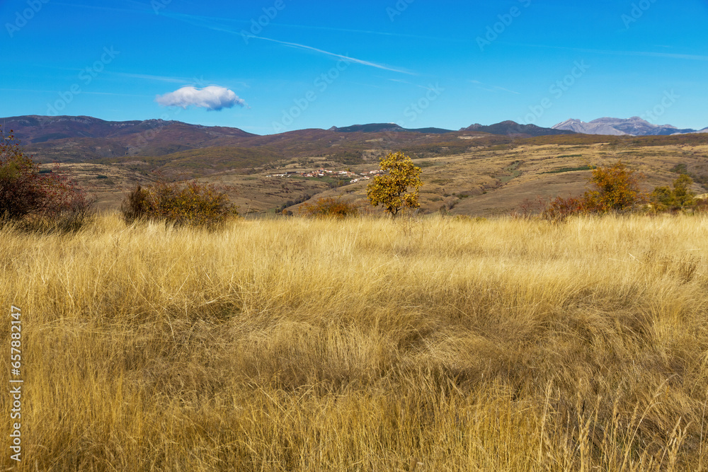 View of rolling autumn landscape with hillside village, dry grass and mountains in the background