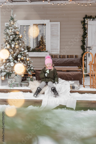 A little girl in winter clothes is sitting on the porch of a house decorated for the Christmas holidays. Christmas holidays for a child.