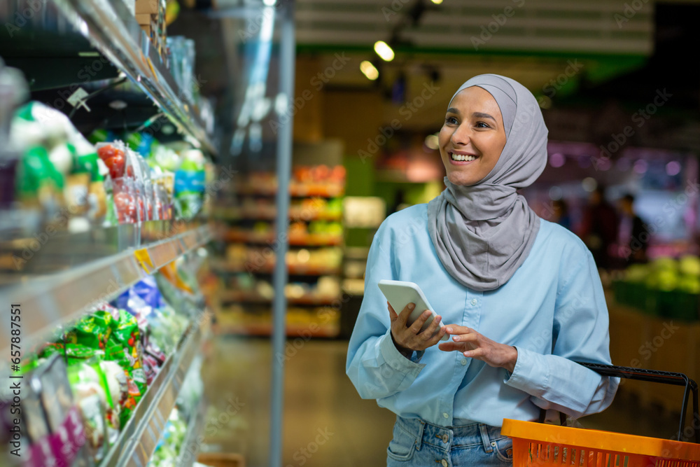Young happy woman shopper with phone chooses products in big grocery store, Muslim woman in hijab uses online shopping list, uses app on smartphone.