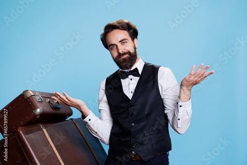 Hotel employee creates i dont know sign, raising shoulders in studio and acting clueless next to luggage. Professional doorman with tie feeling uncertain and doubtful, acting unreliable.