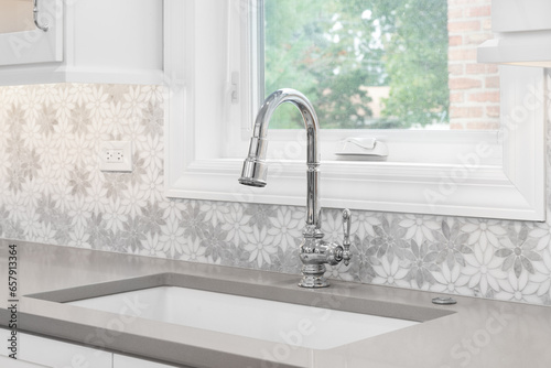 A kitchen faucet detail with a marble daisy flower tiled backsplash, white cabinets, chrome faucet, and a light brown quartz countertop.