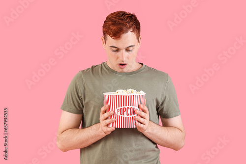 Shocked young redhead man with bucket of popcorn on pink background