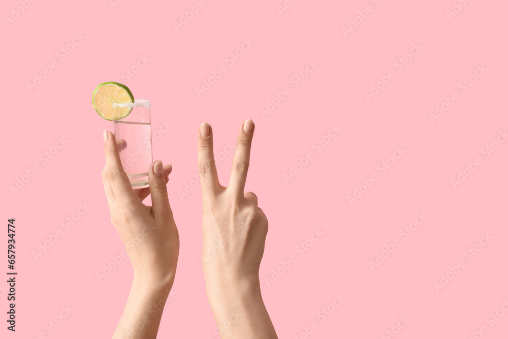 Female hands holding shot of tequila and showing victory gesture on pink background - obrazy, fototapety, plakaty 