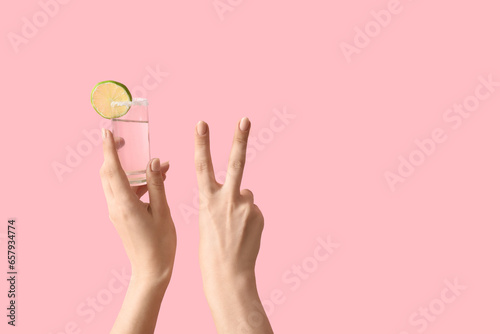 Female hands holding shot of tequila and showing victory gesture on pink background