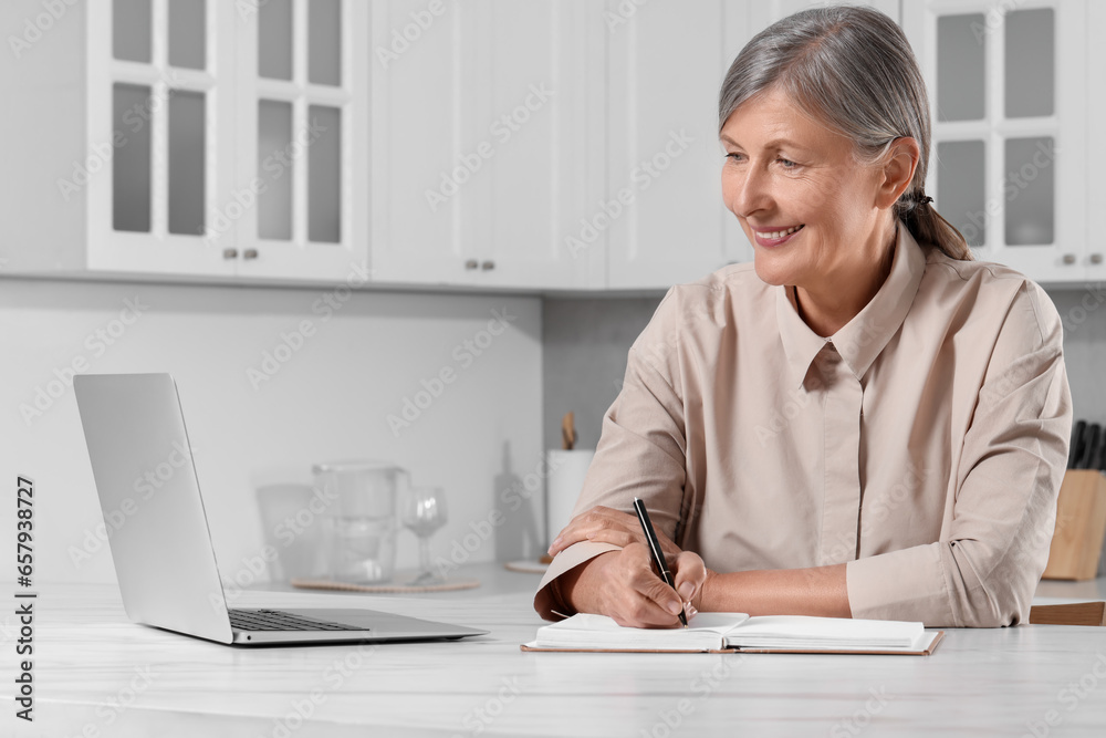 Beautiful senior woman writing something in notebook while using laptop at white marble table indoors Beautiful senior woman writing something in notebook while using laptop at white marble table indoors