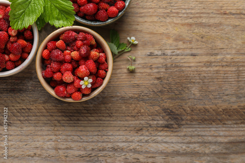Fresh wild strawberries in bowls and leaves on wooden table, flat lay. Space for text