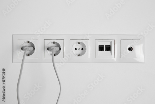 Many power sockets with plugs, ethernet and TV coax plates on white wall indoors photo