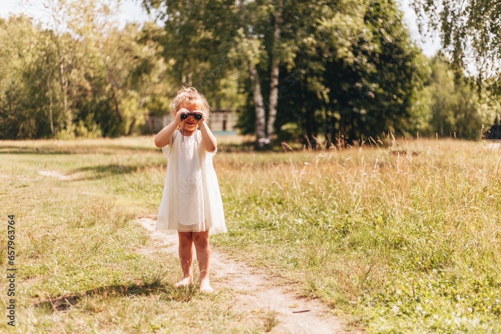 A little blond girl in a white dress is looking through binoculars on a sunny summer day outside
