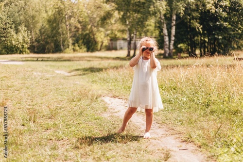 A little blond girl in a white dress is looking through binoculars on a sunny summer day 