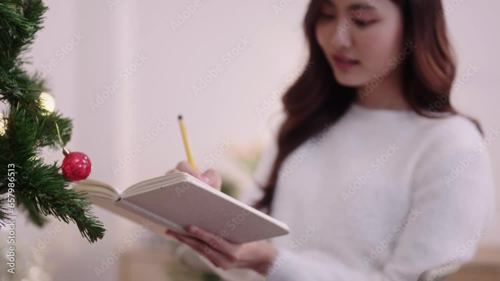 Asian woman sits on sofa near christmas tree tree atmosphere decoration indoors to writing resolutions for New year in notebook at home, winter holidays and people concept Asian woman sits on sofa near christmas tree tree atmosphere decoration indoors to writing resolutions for New year in notebook at home, winter holidays and people concept