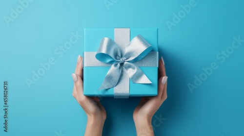 Generative AI Flat lay composition for happy birthday party. Minimalistic greeting card. Women's hands hold a blue gift box with a satin bow on a blue background. Top view.