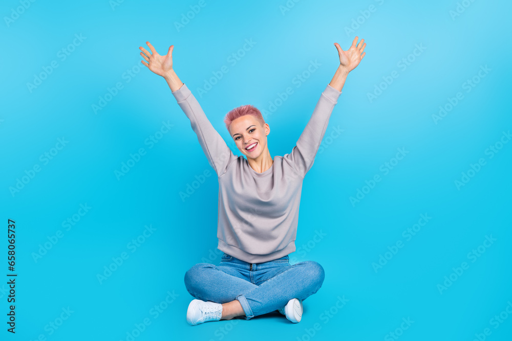 Full length photo of overjoyed cheerful person sit floor crossed legs raise arms isolated on blue color background Full length photo of overjoyed cheerful person sit floor crossed legs raise arms isolated on blue color background