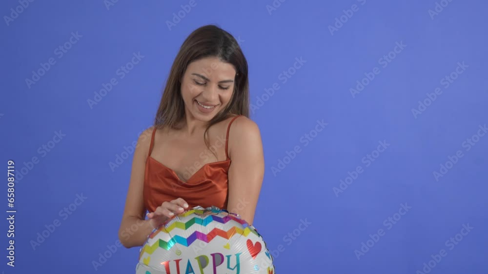 A young woman poses with a Happy Birthday balloon, isolated on a blue studio background. A young woman poses with a Happy Birthday balloon, isolated on a blue studio background.