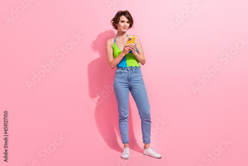 Full length picture of charming young girl with brown bob hair browsing social medias using smartphone isolated on pink color background