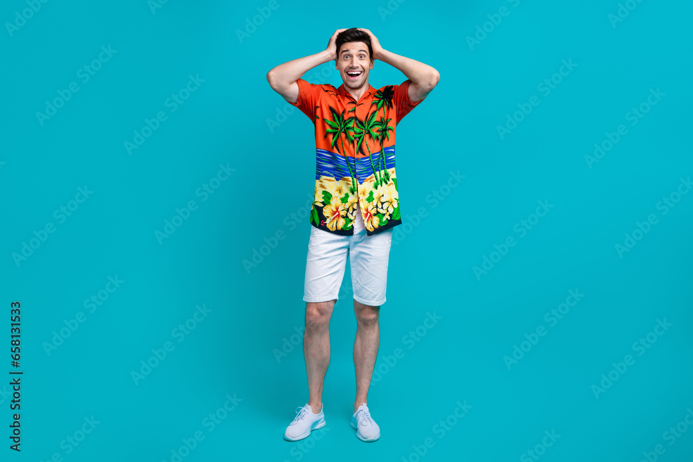 Full size photo of impressed guy wear tropical shirt stylish shorts arms on head astonished staring isolated on blue color background