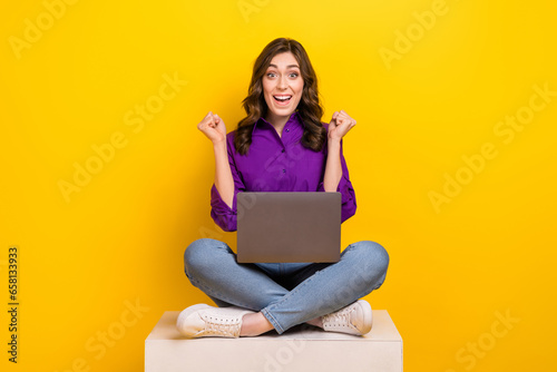 Full length photo of delighted satisfied person sit podium cube use netbook raise fists isolated on yellow color background