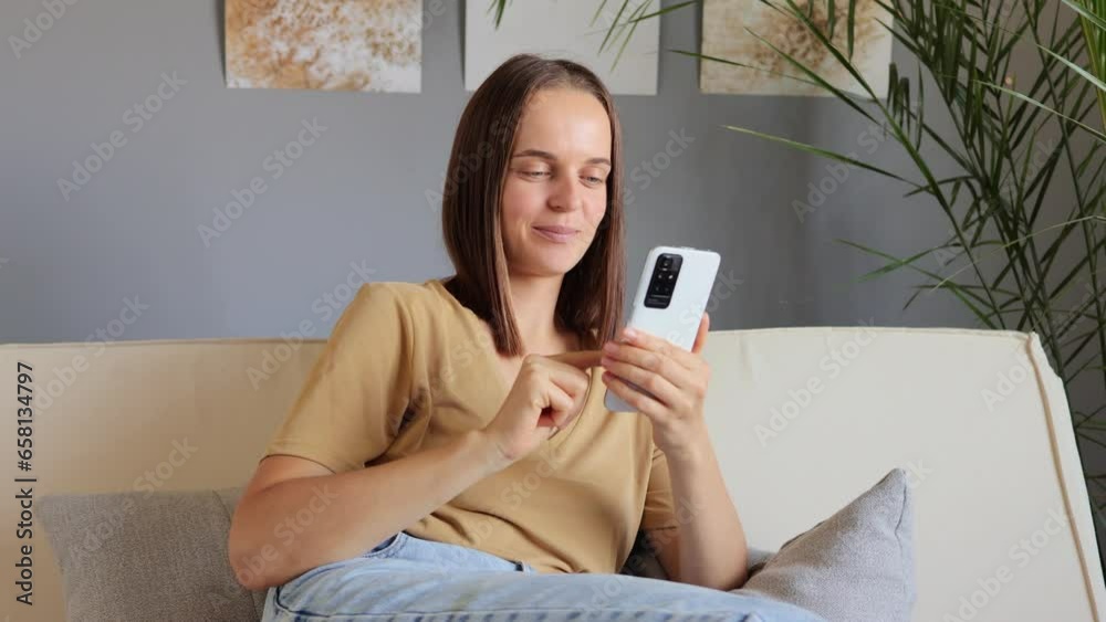 Attractive lively brown haired woman resting on cozy sofa with smartphone sharing messages to friend in social networks smiling enjoying free time on internet scrolling online. Attractive lively brown haired woman resting on cozy sofa with smartphone sharing messages to friend in social networks smiling enjoying free time on internet scrolling online.
