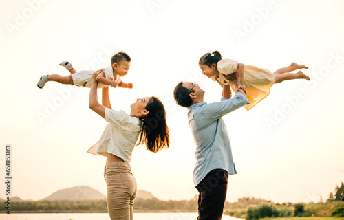 In a sunny park, a father and mother throw their happy son and daughter up in the air, while the children laugh and enjoy the playful freedom of flying. A moment of childhood happiness and family love