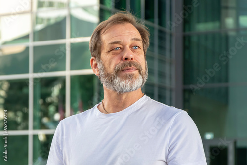 Street portrait of a confident and muscular man 50-55 years old with a gray beard wearing a white T-shirt against the background of a modern glass building. 