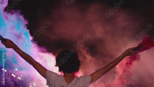 Young woman standing in middle of sport cars and waving , holding colorful smoke bombs flares . Girl starting muscle car drag racing at night . Back view slow motion . Ready for racing . Crowd of fans