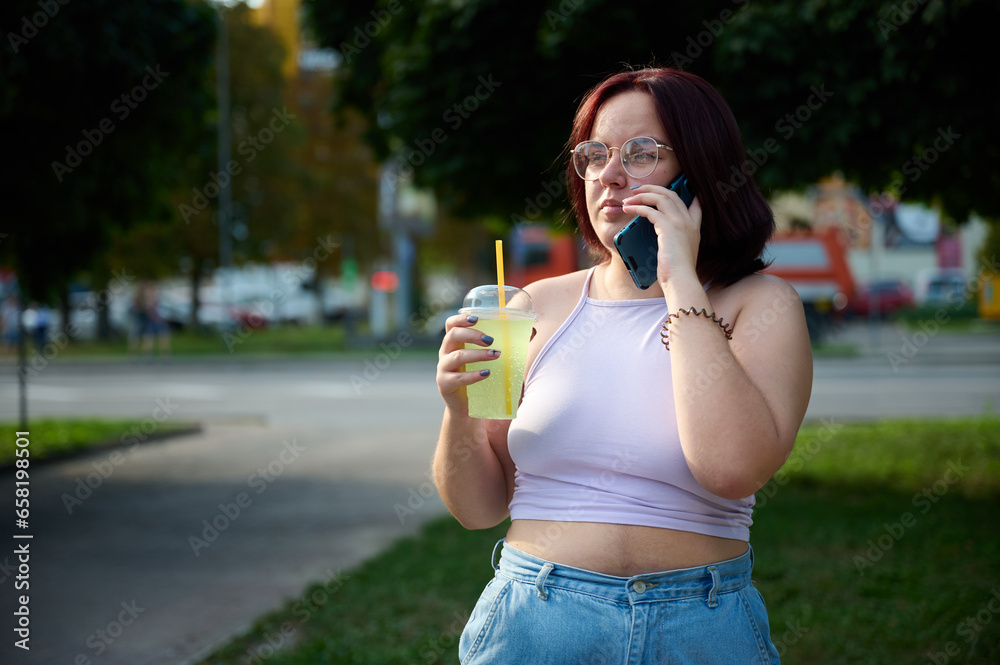 Pretty girl drinking lemonade and talking on phone at sunny day. Young female squinting her eyes because of sunlight. Positive girl drinking and relaxing outside. Pretty girl drinking lemonade and talking on phone at sunny day. Young female squinting her eyes because of sunlight. Positive girl drinking and relaxing outside.