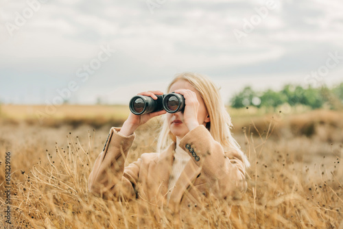 Blond woman looking through binoculars in field under sky photo