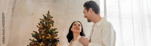 happily married couple in soft home wear decorating Christmas tree in modern apartment, banner © LIGHTFIELD STUDIOS