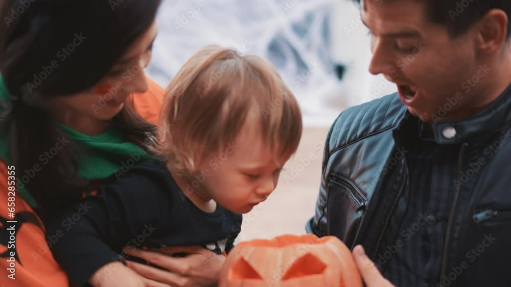 Joyous happy mix race Asian family sitting at decorated porch smiling and laughing. Little boy playing with pumpkin on Halloween. Curious baby blow candles. Dad mother and cute baby celebrate together