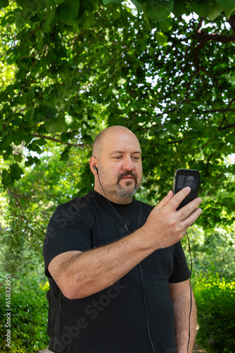 Bald 45-50 year old man with a mustache and goatee wearing a black T-shirt and looking at his cell phone with headphones on in an outdoor park © Beti Argi