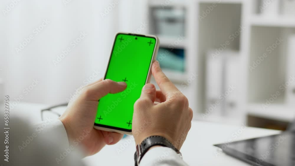 Hospital. Experienced doctor in doctor's clothing holding a smartphone with a green screen in his hands in the office. In motion hospital workers modern phone in hands holding green screen