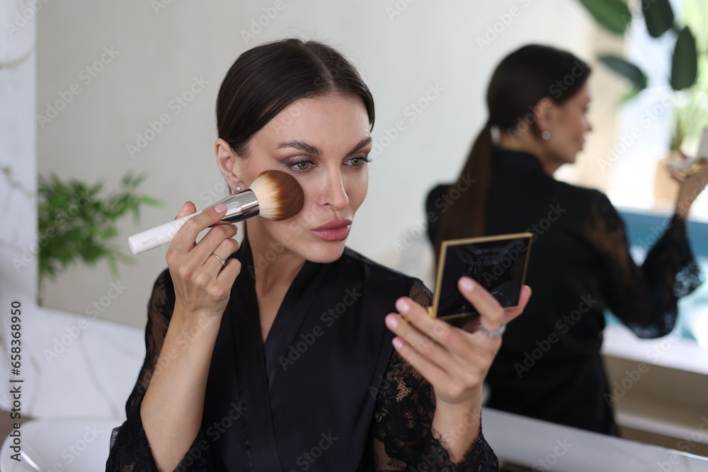 Beautiful brunette woman doing self make-up with a brush and powder. Portrait of young pretty woman in black night gown in her bathroom near mirror. Closeup, big lips. Plants, wooden table. Beautiful brunette woman doing self make-up with a brush and powder. Portrait of young pretty woman in black night gown in her bathroom near mirror. Closeup, big lips. Plants, wooden table.