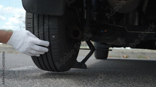 Man turns car wheel on jacked up car and finds screw in tire in city. Puncture of wheel and replacement on asphalt road with protective gloves. Driver finds cause of tyre depressurization. photo