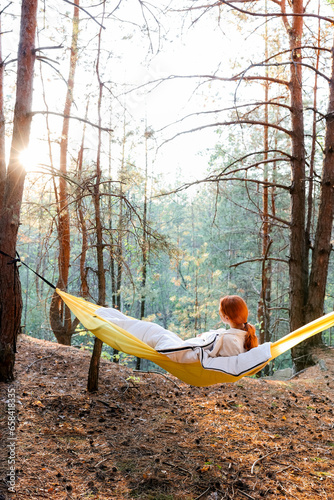 A girl in a hammock in an autumn pine forest looks at the sunset