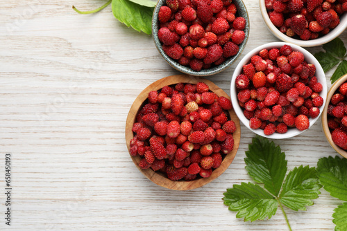 Fresh wild strawberries in bowls and leaves on white wooden table, flat lay. Space for text