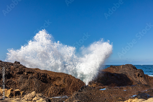 Wave breaking on rock in Point Lobos Nature Preserve, Monterey, California. Spray in the air. Blue sky, Pacific ocean in the background. 