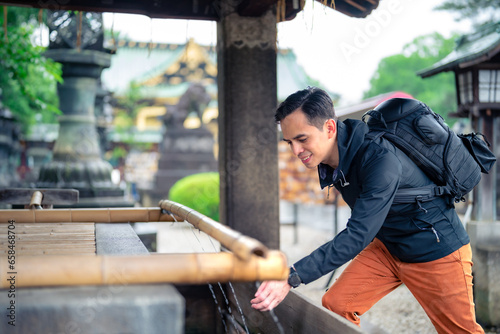 Side view of smiling young Hispanic male tourist backpacker with rucksack touching flowing water in old fountain Kaneiji temple on sunny day in Japan, Tokyo